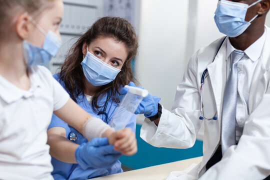 Medical Asisstant Helping African American Pediatrician Doctor To Bandage Broken Arm Of Little Child Patient During Clinical Physiotherapy In Hospital Office. Team With Face Mask Against Coronavirus