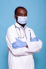 Vertical portrait of confident medic with arms crossed wearing face mask and latex gloves before scrubbing in. Doctor with stethoscope wearing protective medical gear and white lab coat.