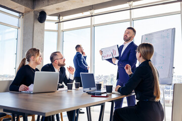 Confident businessman makes a presentation of a new project in the boardroom at a company meeting. Beautiful auditors talk with different partners about the business using a whiteboard and graphs.