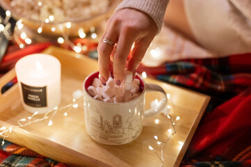 woman's hand applies marshmallows on cup of cocoa on tray on red plaid and garland of glowing lights at mobile home trailer. concept taking care of yourself and relaxing on new year and christmas eve