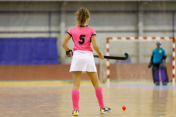 Young female indoor hockey player standing with the ball across goalkeeper.