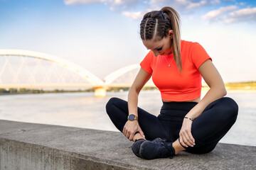 Young attractive fitness girl in black yoga pants and orange short shirt sits and relax her body in lotus posture face downwards on short concrete wall near river during the day.
