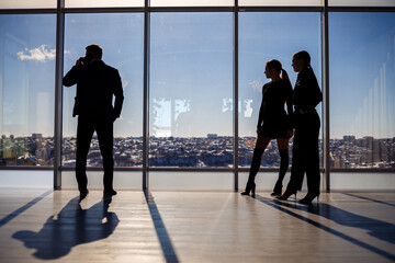 Two women and a man in the office enjoy the view of the city from the window of a skyscraper and talk while standing by the large window in the office