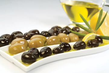Glass bottle of homemade olive oil and olive tree branch, raw turkish green and black olive seeds and leaves on white table.