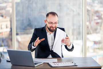 Fototapeta premium Adult male businessman, teacher, mentor is working on a new project in the office and is nervous. Sits at the table by the large window. He works on a laptop, business problems