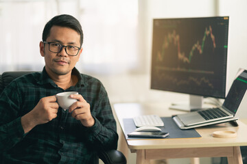 Asian man trader drinking coffee and sitting at home office in front of monitors with...