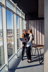 Two successful female architects are talking about a joint project while standing at the window with a laptop. Young women economists dressed in formal attire talking during a break from work