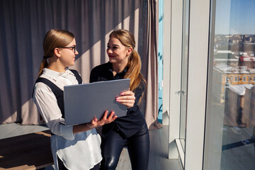 Two successful female architects are talking about a joint project while standing at the window with a laptop. Young women economists dressed in formal attire talking during a break from work