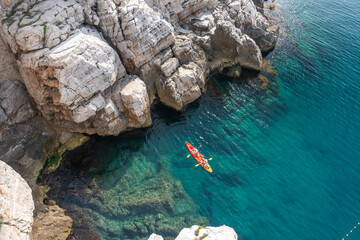 View from the rock cliffs of kayaker exploring the crystal clear Mediterranean waters of a cove off the coast of Dubrovnik, Croatia - obrazy, fototapety, plakaty