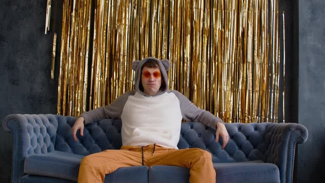 Serious and dissatisfied man in hood with ears and bright glasses sitting on sofa against wall with golden tinsel. Relaxed important male sprawled on couch and looking at camera.