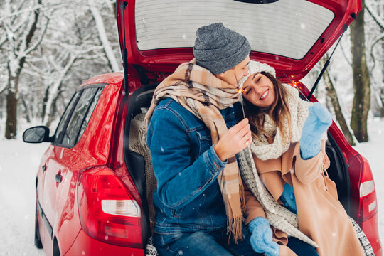 Young couple in love burning sparklers in car trunk in snowy winter forest. People relaxing outdoors during road trip