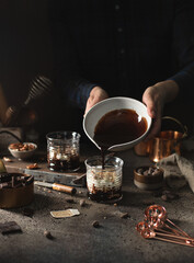 Women pours hot chocolate in cup with curd dessert with various copper dishes,chocolate beans and wooden dishes on dark background.