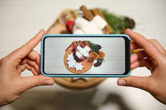 Phone in real time. Close-up. Hands holding phone and taking pictures of gingerbreads in the shape of stars laid out on wooden board and on baking paper next to dried orange slices, pine cones, twigs