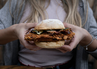 Eating. Closeup view of a young woman holding a crispy fried chicken sandwich with bacon and sliced cucumber.