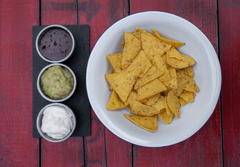 Mexican food. Top view of nachos in a white bowl and three dipping sauces of cheddar cheese, sweet chili and guacamole, on the red wooden table. 