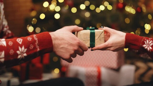 Close-up hands of man giving festive box with Christmas present to woman sitting at dinner feast table during holiday family party, decorating xmas tree and celebration lights, selective focus