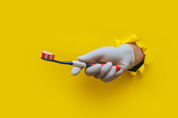 Doctor's hand in a white latex medical glove holds a toothbrush with toothpaste. Torn hole in yellow paper. Dental care and oral hygiene concept.
