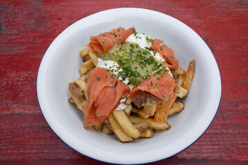Top view of french fries with smoked salmon, cream cheese and guacamole sauce in a white bowl on the red wooden table.
