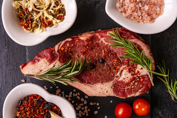 Ribeye raw piece of beef fillet on a slate board and spices for meat rosemary, pink salt and pink pepper. Cooking a dish. Everything for a barbecue.