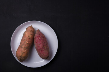 Orange, purple raw sweet potatoes, yam on white plate, black table. Farm harvest, organic vegetables, shop local vegan food. Top view, copy space