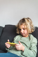 Naklejka na ściany i meble Cute little kid with blond hair in casual clothes resting on cozy couch and playing video game on smartphone during weekend at home