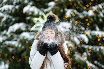 young woman blows snow from her hands while standing against the background of a Christmas tree