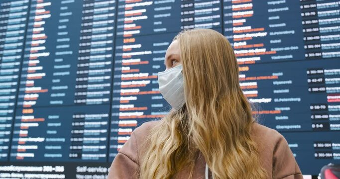 Young woman travelling with face mask to protect from COVID-19. Passenger beside scoreboard in airport with flights schedule blurred