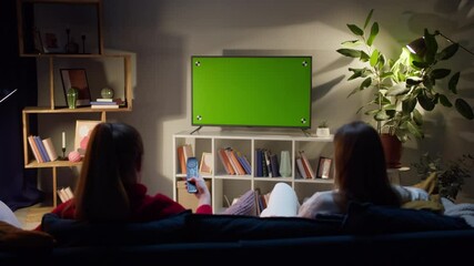 Women watching television with chroma green screen. Girlfriends switching channels on tv with remote controller, sitting on sofa in living room. Spare time at home in evening.