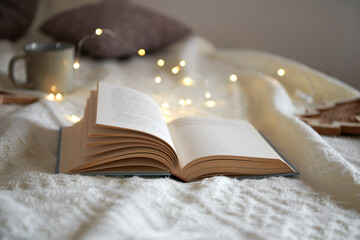 Opened book on a knited beige plaid, christmas garland lights and mug, cozy winter day at home....