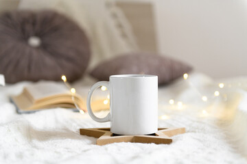 White mug mockup with blank copy space. Coffee cup on a beige knited background with opened book and christmas garland lights, cozy winter day at hugge home