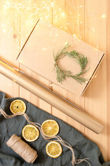 Fototapeta na wymiar Idea of eco-friendly packaging for Christmas gifts. Box, whip, wreath of green twigs, dried orange slices, paper, muslin cloth, garland with light on wooden table. Step-by-step instruction. DIY. Step1