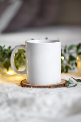 Obraz na płótnie Canvas White mug mockup with blank copy space. Coffee cup on a beige knited background with eucaliptus leaves and christmas garland lights, concept Christmas cozy winter day at hugge home, selecive focus