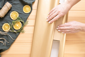 Female hands are wrapping a gift box in paper. Wooden table on the background. Dried orange slices on a green muslin cloth.Top view. DIY concept. Step-by-step instruction. Step 2.