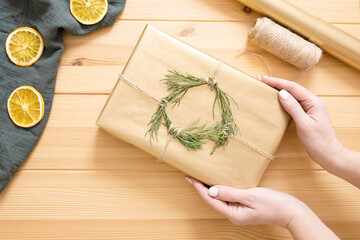 Idea of eco-friendly decor for Christmas gifts. Women's hands are holding beautifully wrapped gift with wreath of green sprigs. Dried orange slices, muslin cloth on wooden table. Top view. DIY. Step 7