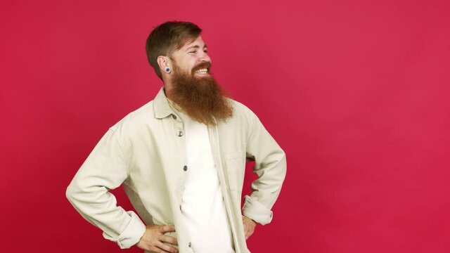 Redhead man with long beard posing with arms at hip and laughing over isolated background