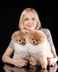 Smiling pretty woman holding two cute puppies of pomeranian spitz breed dog. Owner with her friends pets at black background.