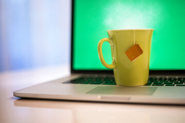 Closeup view of a cup of hot beverage and a laptop with green screen in background. Steaming yellow mug on computer keyboard. Remote workstation. Smart working, home working and homeschooling concept