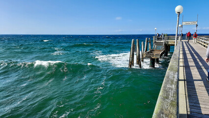 Fototapeta premium Steg des Ostseebad Wustrow . Türkis blaues Wasser mit Wellen und einigen Touristen auf dem Holzsteg . Schild Ostseebad Wustrow bei leichtem Wellengang und strahlend blauem Himmel . East Germany Marine