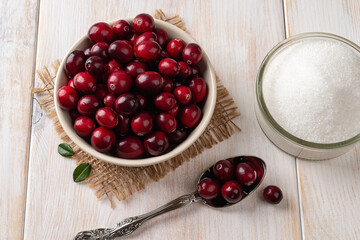 Fresh red cranberries in a beige bowl, spoon and sugar over white rustic wooden table. Ingredient for cooking dried cranberries, Thanksgiving and Christmas dishes. Wild berries for healthy eating.