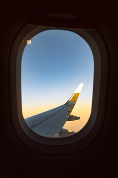 Fotograf&iacute;a en formato vertical de la ventanilla de un avi&oacute;n con el ala de fondo, con una franja amarilla al amanecer.