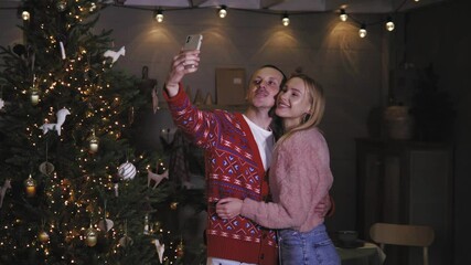 Happy caucasian couple taking selfie photo in front of christmas tree celebrating christmas night together, sending congratulation on mobile phone smartphone, having video call conference 