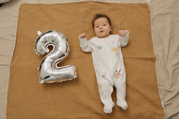 An infant is relaxing on a muslin blanket near a silver foil balloon in the shape of 2. A baby girl in one-piece clothing is celebrating her two months.