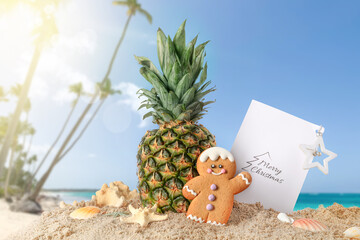 Christmas composition with pineapple, gingerbread cookie and greeting card on sand at tropical...