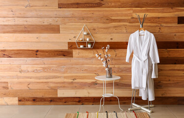 Cotton branches in vase on table and rack with bathrobes near wooden wall - obrazy, fototapety, plakaty