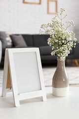 Vase with gypsophila flowers and blank frame on table in living room