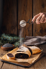 Woman sprinkling sugar powder onto tasty poppy seed strudel on wooden background