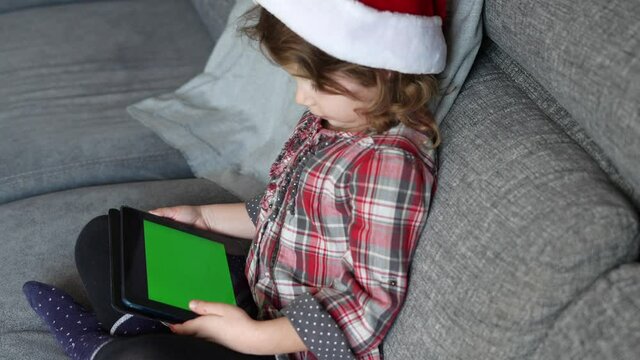 Little girl sitting on the sofa at home with Santa Claus hat holding and looking at tablet with green screen. Kids and gadjets concept