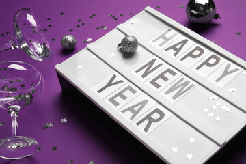Board with text HAPPY NEW YEAR, glasses and Christmas decor on purple background, closeup