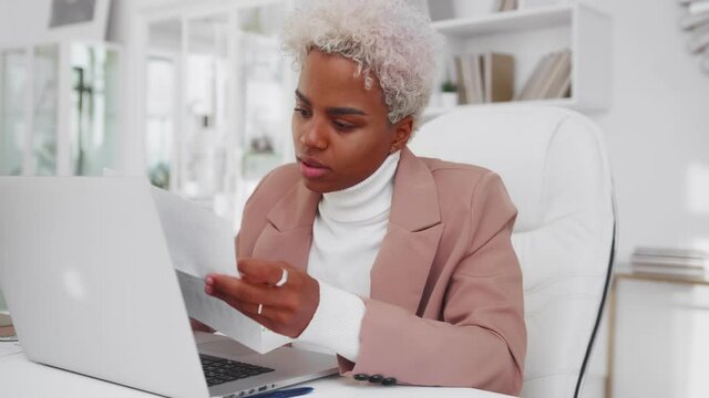 Puzzled young African American woman entrepreneur read letter makes frightened look dissatisfied with seeing correspondence and bad news, received incorrect data sits at office table with laptop.