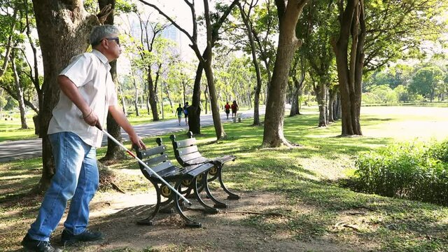 The life of an Asian senior blind man uses a cane to guide him and sits in a wooden chair to sit and relax in the afternoon garden in the clear air.